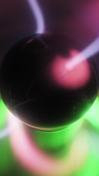 Vertical close-up of a plasma globe with pink and purple electric bolts moving from a central electrode. Ionized gas tendrils flash over a glowing green base.