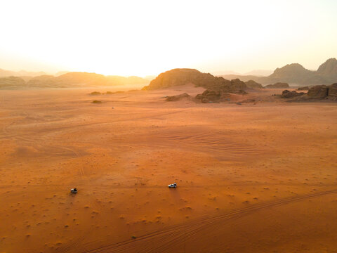 Aerial view of Wadi Rum desert landscape with red sand dunes, rocky mountains, and off-road vehicles driving across the vast terrain during sunset, Wadi Rum, Aqaba Governorate, Jordan.