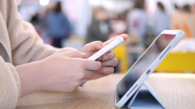 Close-up of hands using smartphone and tablet. Person focused, calm, multitasking in public space. Ideal for tech, education, or digital lifestyle themes. Blurred background suggests busy environment