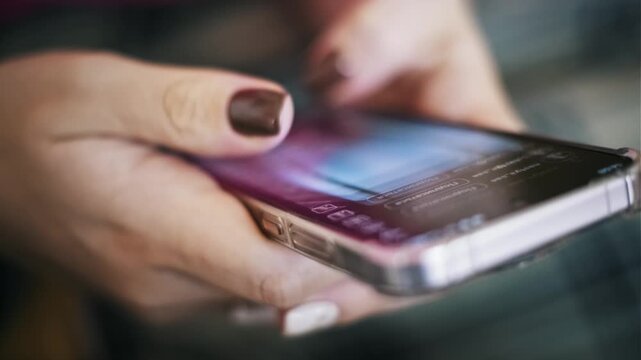 Side angle of female hands with a brown manicure scrolling a smartphone screen. The thumb swipes the touch display of the device in a clear case against a blurry background.