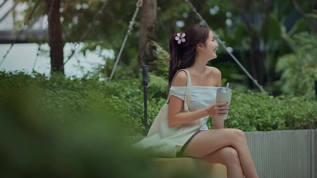 Happy Asian woman smiling and holding reusable tumbler while sitting at green public park, beautiful young female traveler carrying tote bag and relaxing outdoors for zero waste, eco friendly.