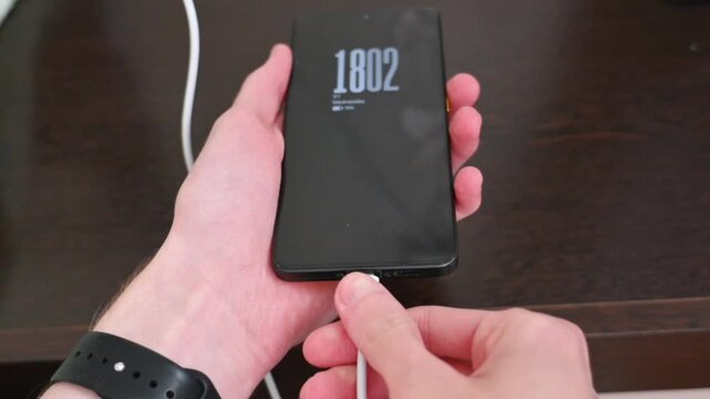 Human hand holding black smartphone with charging cable on wooden desk close up