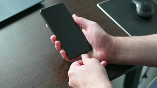 Human hand holding black smartphone with charging cable on wooden desk close up