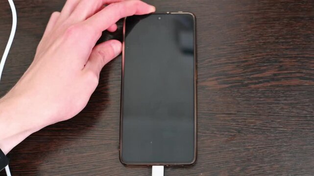Human hand holding black smartphone with charging cable on wooden desk close up