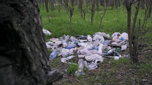 A large pile of plastic bottles and bags lies on the grass in a deciduous forest. The camera slides past a tree trunk to reveal the extent of environmental pollution.
