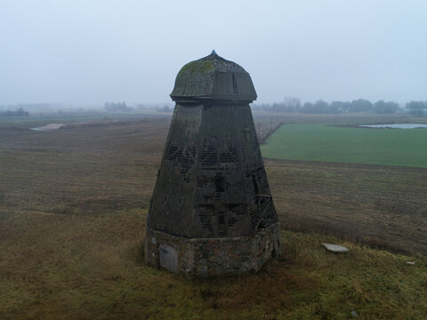 Aerial view of an old dilapidated wooden windmill standing in a vast rural field under a foggy sky in Kakiske, South Kurzeme Municipality, Latvia.