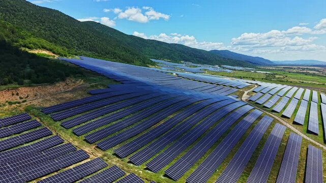 Solar panels are arranged in rows across a valley with green hills and mountains in the background on a sunny day. The blue sky has a few clouds, and the landscape is bright.