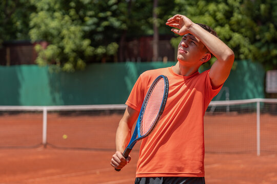 Junior tennis player squinting in bright summer sun during outdoor practice on clay court. Summer sports concept highlighting sun exposure, UV protection, heat and outdoor safety