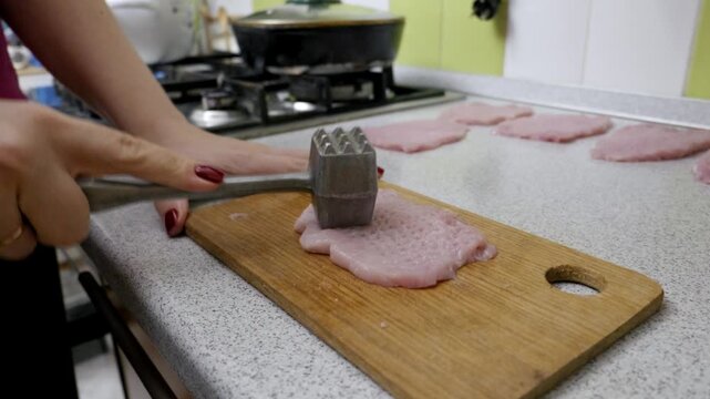 Woman's hands use a metal meat mallet to pound and tenderize a raw pork cutlet on a wooden cutting board. Close-up shot captures the food preparation process in a domestic kitchen.