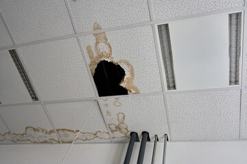 Fototapeta premium Water damage on a suspended office ceiling with a large hole and brown stains from a leak. Damaged tile in a T-bar ceiling structure requiring repair due to flooding or pipe burst.