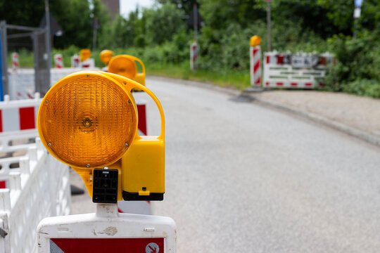 Yellow construction warning light mounted on a white barricade, with a blurred road and green vegetation in the background indicating a construction zone