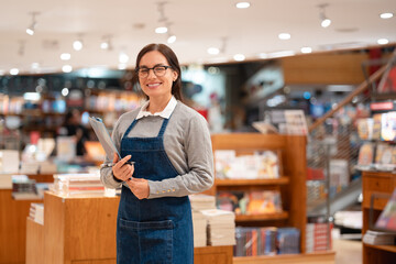 Fototapeta premium Happy Latin Brazilian Manager with Glasses in Gray Sweater and Apron Holding Clipboard in Bookstore Wide Medium Shot