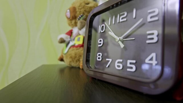 Modern analog alarm clock on a bedside table shows time passing quickly in a timelapse. A soft-focus stuffed toy sits in the background against a green wall.