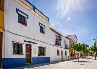 Fototapeta premium White residential houses with traditional blue accents along a sunny paved street, featuring potted flowers in windows and small trees under a clear blue sky in Denia, Costa Blanca