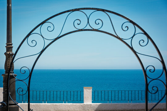 Wrought iron arch featuring decorative scrolls framing the expansive blue Mediterranean Sea and clear sky from a viewpoint in Altea, Alicante, Spain, providing a serene and scenic outlook