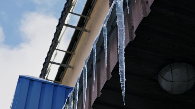 Row of sharp, frozen icicles hangs from the edge of a dark roof eave. A low-angle shot tilts up, revealing a bright blue sky with white clouds in the background.