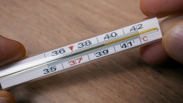 Fingers hold a glass clinical thermometer with a rising temperature indicating a high fever. Close-up shows the Celsius scale as the liquid column rises rapidly past 42 degrees against a