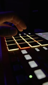 A musician's hand taps rhythmic beats on illuminated MPC drum pads in a dark studio setting. Concept of music production, electronic creative process, and nightlife entertainment.