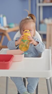 Toddler girl drinking from yellow sippy cup, holds cup to mouth in kitchen highchair with bowls and spoon on tray; calm domestic morning.