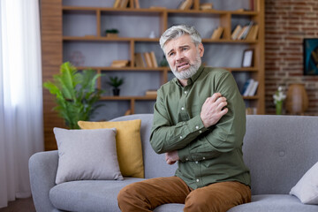 Fototapeta premium Mature man sitting uncomfortable on a sofa at home, scratching his arm and looking distressed, suffering from an allergic reaction or skin condition symptoms