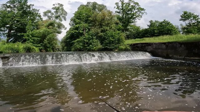 POV of Fisherman Casting Rod near a River Weir with Flowing Water and Green Broadleaf Trees (Magnoliophyta), Summer Angling.