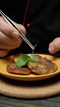 Chef carefully garnishes fried plantains on a yellow plate with fresh green leaves using tweezers in a well-lit kitchen setting