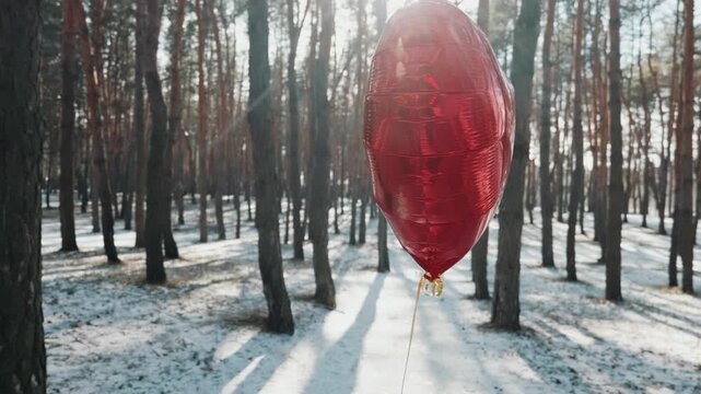 Red foil heart balloon floats in a winter pine forest covered in snow as sunlight shines through trees. Concept illustrates a romantic Valentine's Day surprise outdoor.