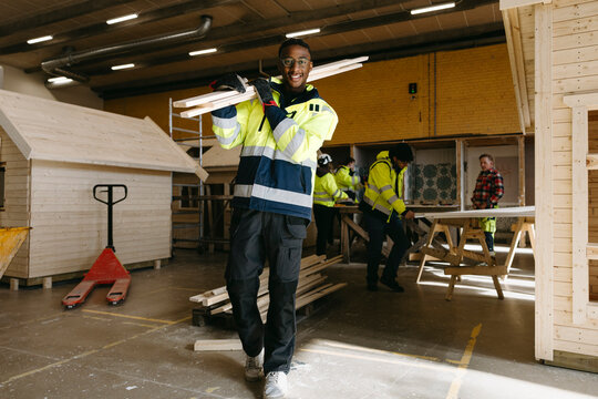 Portrait of smiling young male student carrying wooden planks on shoulder in vocational workshop