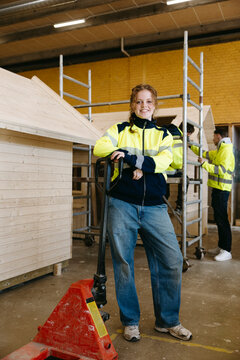 Portrait of smiling young female student wearing safety jacket standing beside red pallet jack in vocational workshop