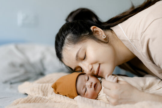 Young mother gently kisses sleeping newborn baby on bed in soft morning light
