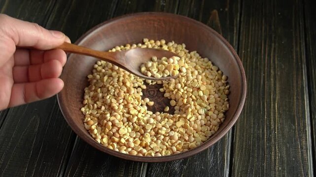 Hand using wooden spoon to stir yellow split peas in a round wooden bowl on a rustic wooden table, showcasing the texture and color of the peas