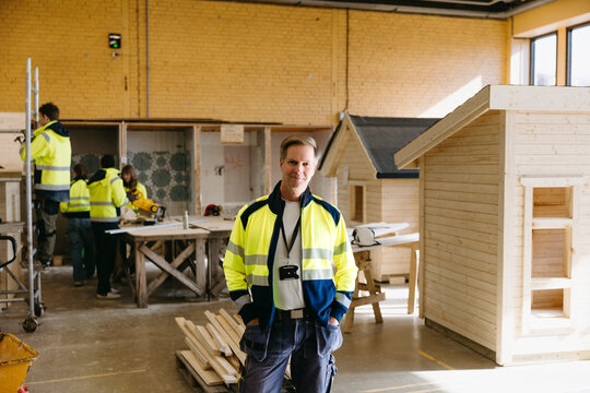 Smiling portrait of mature male instructor standing with hands in pockets at construction workshop