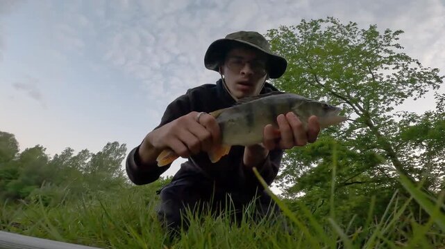 Proud fisherman in bucket hat showing a caught European perch (Perca fluviatilis) to the camera. Low angle outdoor freshwater fishing success.