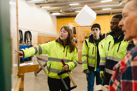 Female student holding clamp points while explaining ideas to attentive classmates on whiteboard inside construction wor