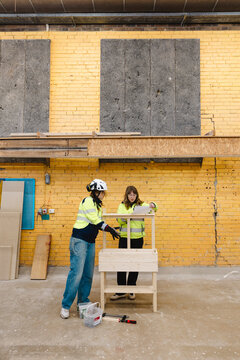 Female vocational students examining blueprint while constructing wooden planter stand against textured yellow brick wal