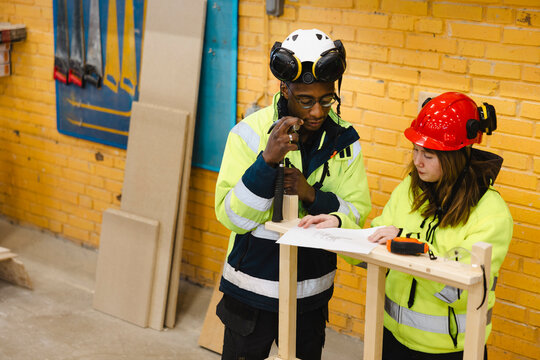 Two vocational students wearing reflective jackets and protective helmets reading blueprint on wooden framing assembly a