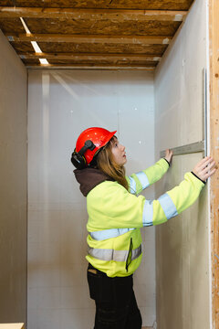 Female vocational student wearing reflective jacket and hardhat measuring drywall panel with ruler inside narrow constru