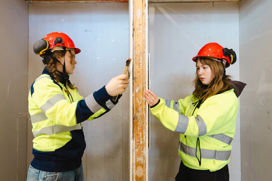 Two female vocational students working together while measuring drywall panel with ruler on opposite sides inside traini