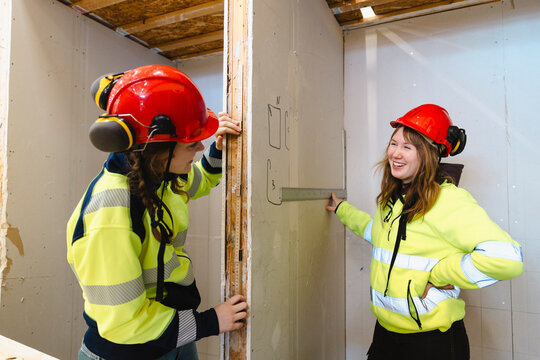 Female student talking with classmate measuring drywall with ruler in construction booth at secondary school