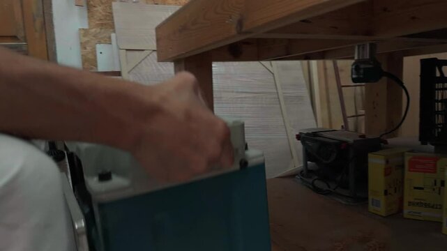 Unrecognizable carpenter in overalls lifting a heavy thickness planer machine from under the workbench and placing it on a wooden table to prepare for timber milling in a local carpentry shop