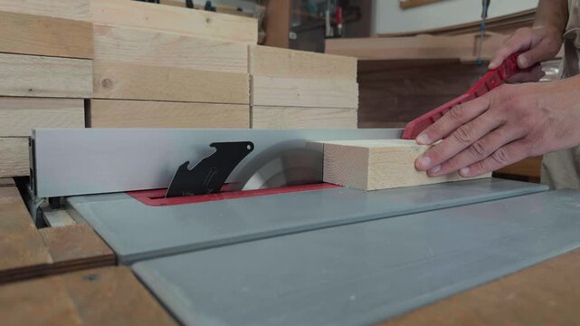 Unrecognizable male artisan hands using a red safety stick to carefully slide a raw timber chunk into a spinning circular saw blade inside a bright maker studio extreme macro push in shot