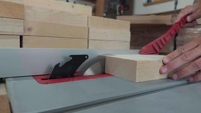 Unrecognizable male joiner hands using a red plastic push stick to safely feed a softwood block through a spinning table saw blade inside a woodworking station close up handheld documentary