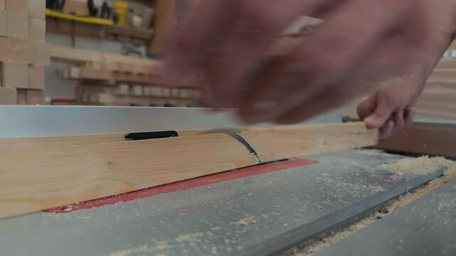 Extreme close up of an unrecognizable joiner using a safety push stick to feed a pine board through a fast spinning circular table saw blade removing a thin edge in a dusty woodworking shop