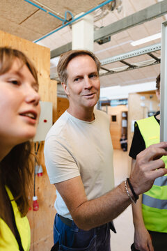 Smiling male instructor explaining components to control panel to students in workshop at vocational school
