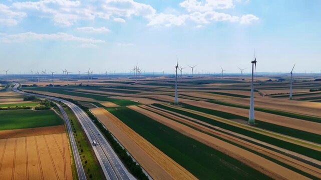 Wind turbine beside road, patchwork farmland. Turbine near paved road, green and brown fields, aerial drone shot, sunny