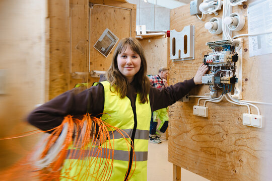 Portrait of smiling vocational student holding bundle of orange wiring while leaning on wall mounted electrical breaker
