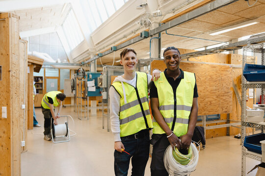 Portrait of smiling young man standing with male friend holding bundle of wires in vocational engineering workshop