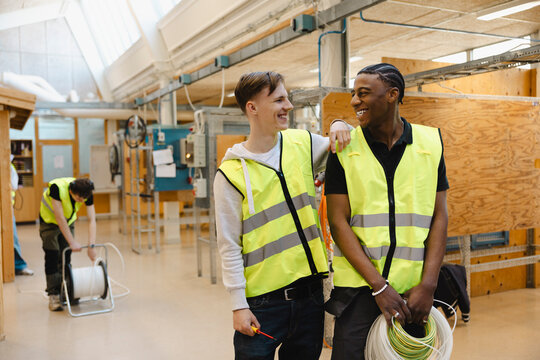 Smiling young man standing with male friend holding bundle of wires in vocational engineering workshop