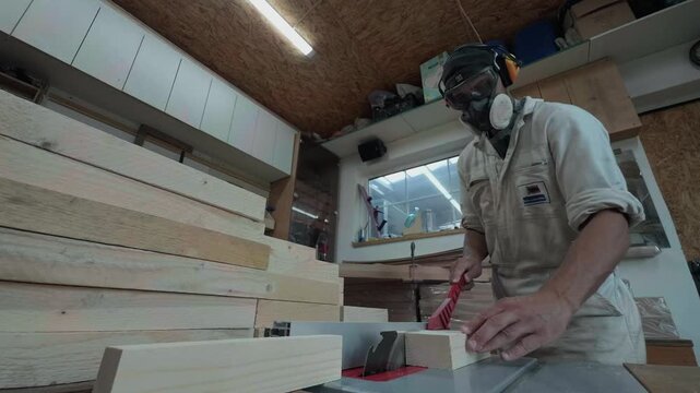 Anonymous male carpenter in safety earmuffs and respirator guiding thick lumber through a spinning circular table saw with a red pusher inside a busy facility static low angle bench level shot