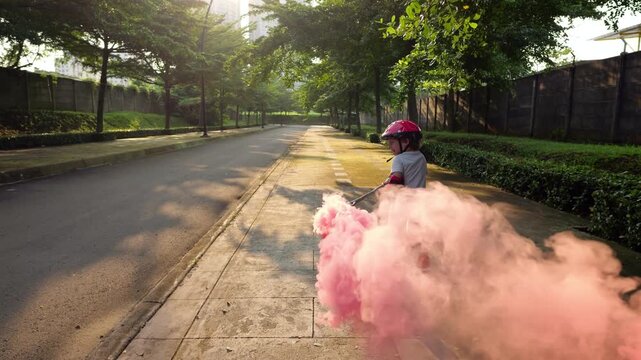 Little boy rollerblading on sidewalk creating bright pink smoke curling in sunlight, camera follows him, slow motion. Young child is not very confident in rollerblading, but is happy with smoke effect